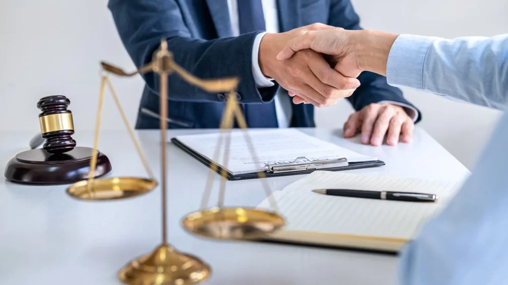 Two people shaking hands over legal documents, with scales of justice in the foreground.